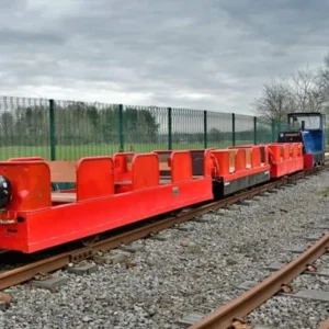 Red gloss-painted flatbed rail wagons on a narrow-gauge track beside a fenced field under a cloudy sky, with a small blue locomotive at the rear.